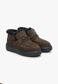 Brown suede ankle boots with a fluffy black lining, featuring a decorative black buckle and a thick black sole. Textured upper with smooth accents.