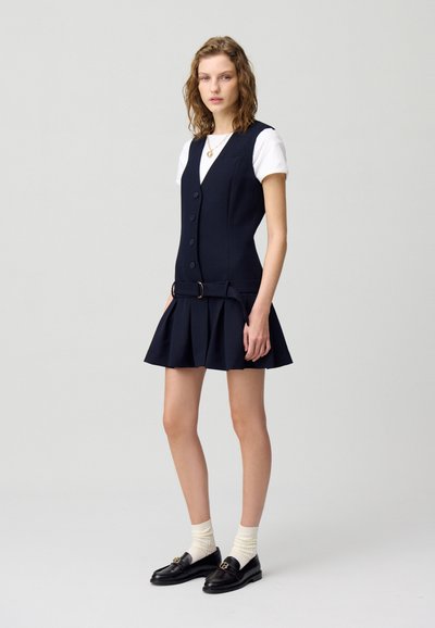 Robe plissée marine avec un bodice ajusté, des boutons sur le devant et un détail de ceinture. Associée à un t-shirt blanc et des mocassins noirs avec des chaussettes.