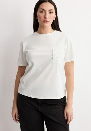 Žena s dlhými tmavými vlasmi, oblečená v bielej košeli s krátkymi rukávmi s čipkovaným vreckom a čiernych nohaviciach, stojaca pred jednoduchou stenou.