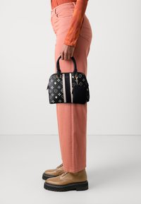 Sac à main structuré noir avec des motifs d'embl�èmes blancs, courtes poignées, détail de rayure rose et une petite poche attachée, tenu contre un pantalon rose.