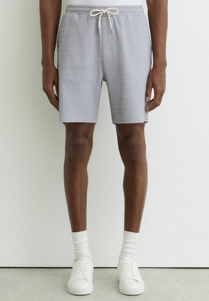 Mand iført lysegrå strikkede shorts med snøre, hvide crew-strømper og hvide lave sneakers mod en ensfarvet baggrund.