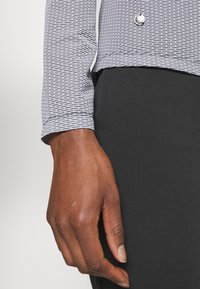 Mörkhyad hand bredvid svarta byxor och en vit långärmad skjorta med ett litet mönster av svarta streck samt en silverknapp.