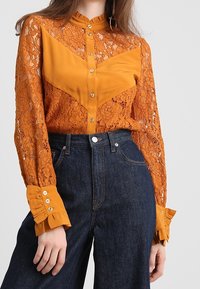 Camicia arancione in pizzo con design a zig zag in tessuto solido, chiusura con bottoni davanti e polsini arricciati, abbinata a jeans a vita alta blu scuro.