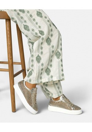 Jambes portant un pantalon blanc avec des motifs verts, assises sur un tabouret en bois, montrant des baskets cloutées dorées avec des semelles blanches.