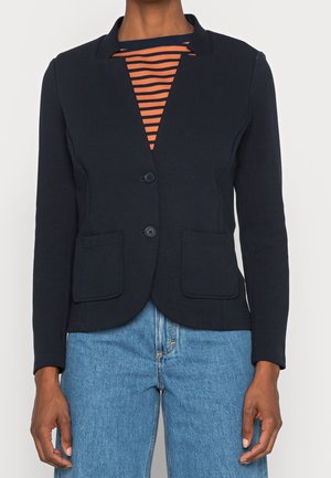 Kobieta w dopasowanym czarnym żakiecie na pomarańczowo-czarnym paskowanym topie, w niebieskich dżinsach, stojąca na jednolitym tle.