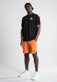 Black short-sleeve t-shirt with a small red and blue logo, paired with bright orange shorts and white sneakers, set against a neutral background.