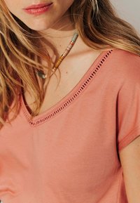 T-shirt couleur pêche avec un décolleté en V et des découpes décoratives. Le tissu semble doux et léger. Un collier en perles met en valeur le décolleté.