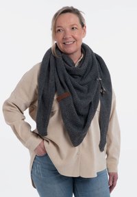 Szary teksturowany trójkątny szal z skórzaną metką, zarzucony na osobę w beżowej koszuli zapinanej na guziki, noszącą niebieskie dżinsy.