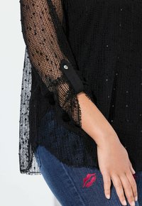 Top nero trasparente con rete testurizzata, decorato con sottili paillettes. Presenta maniche lunghe con polsini bottonati. Indossato sopra denim con stampa di rossetto.