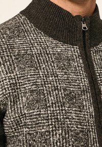 Primo piano di un maglione a maglia a scacchi marrone e beige con colletto a coste e una zip metallica a metà.