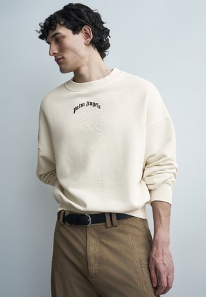 Νεαρός άνδρας με σγουρά μαλλιά που φοράει μια κρεμ φούτερ Palm Angels και καφέ παντελόνια, κοιτάζοντας προς τα πλάγια μπροστά σε απλό φόντο.