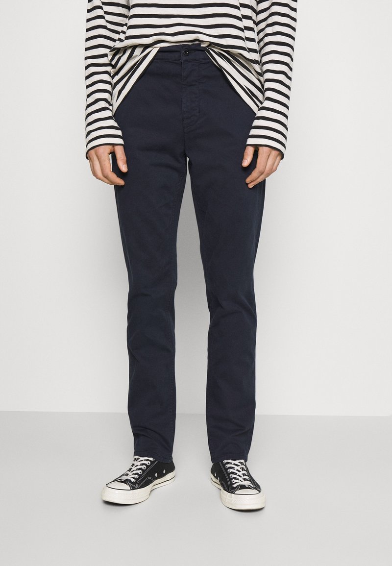 Personne portant des baskets basses noires, un pantalon droit bleu marine foncé, et une chemise blanche à manches longues avec des rayures horizontales noires.