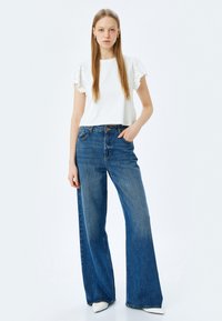 Witte top met korte mouwen en gerimpelde randen, gecombineerd met hoge taille, wijde blauwe jeans. Model staat voor een effen achtergrond.