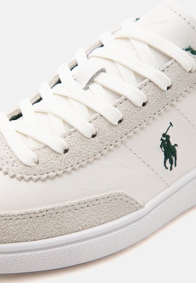 Gros plan d'une basket blanche avec garniture en daim, lacets blancs, semelle texturée et logo brodé représentant un joueur de polo vert foncé sur le côté.