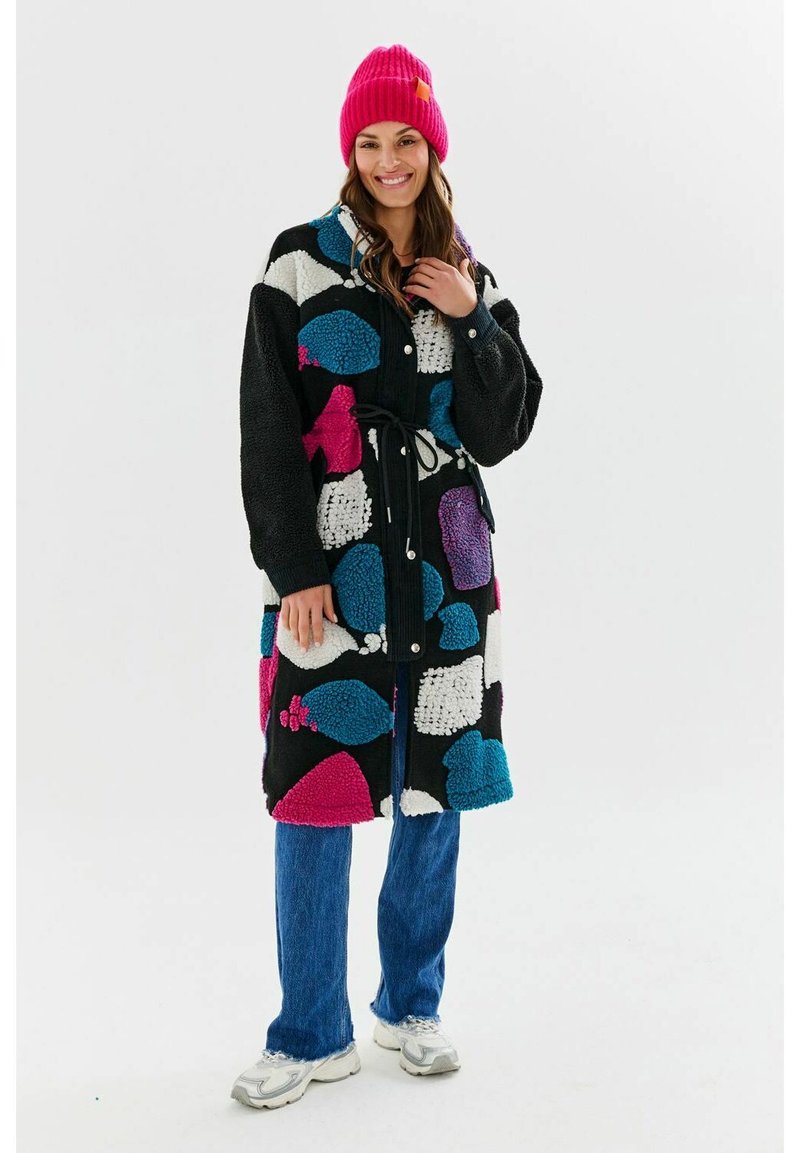 Kolorowa, wzorzysta odzież wierzchnia z dużymi różowymi, niebieskimi i białymi kształtami na czarnym tle, z paskiem i guzikami. Zestawiona z niebieskimi dżinsami.