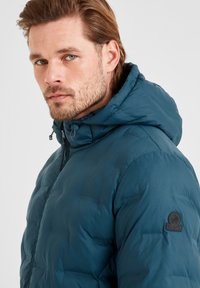 Teal puffer jacket with a hood featuring a zip closure, textured fabric, and a logo badge on the left shoulder.