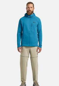 Giacca blu con cappuccio in tessuto testurizzato e tasca con zip, abbinata a pantaloni beige chiaro rimovibili con cuciture decorative e vestibilità dritta.