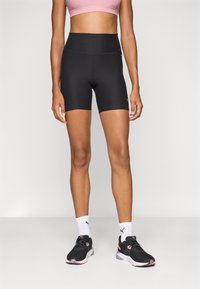 Hoch taillierte schwarze Radshorts aus dehnbarem Stoff, akzentuiert mit flachen Nähten, kombiniert mit schwarzen Sneakers und weißen Socken.