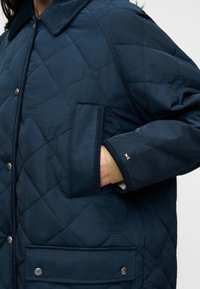 Ναυτικό μπλε καπιτονέ μπουφάν με γιακά από κορδόνι, κουμπιά με πατιλέτα, πλαϊνές τσέπες και ριμπ μανσέτες. Διαθέτει οριζόντια ραφή διακοσμητικά στοιχεία.