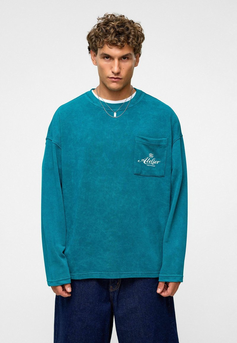 Teal lange mouwen shirt met een borstzak, zachte textuur, oversized pasvorm en een wit logo. Gecombineerd met losse, donkere jeans.