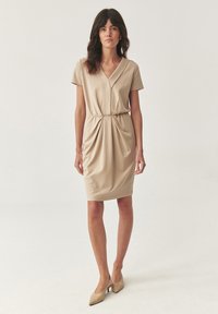 Abito beige all'altezza del ginocchio con maniche corte, scollatura a V e dettaglio in vita arricciata. Tessuto liscio e elasticizzato con una texture morbida.