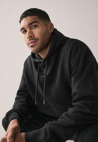 Sudadera con capucha de algodón negra con cordón ajustable en la capucha, corte holgado, puños acanalados y detalle de costura plana. Modelo sentado sobre un fondo neutro.