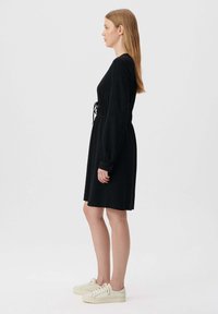 Schwarzes langärmliges Kleid mit geraffter Taille, texturiertem Stoff, knielangem Schnitt und geknöpften Manschetten, kombiniert mit hellen Sneakers.