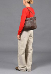 Bolso de hombro tejido en marrón con un acabado suave, que presenta una sola correa y una forma rectangular, llevado sobre una blusa roja y pantalones beige.