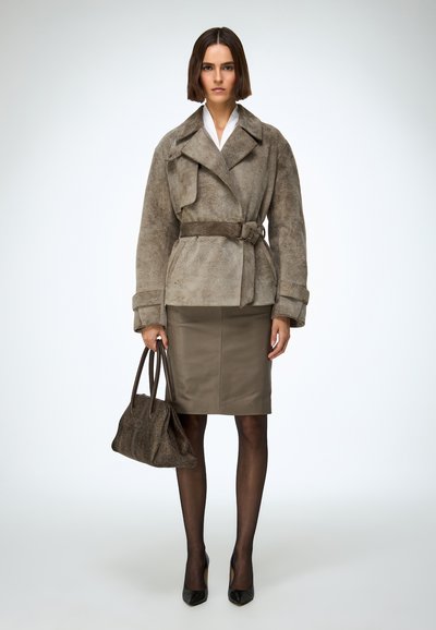 Barna szarvasbőr wrap kabát övvel a derekán, olivazöld testhezálló szoknyával párosítva. Fekete magassarkú cipő és texturált kézitáska egészíti ki az összeállítást.