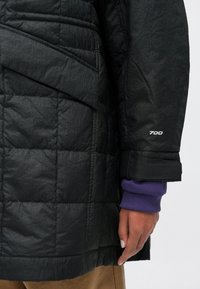 Czarna pikowana kurtka puchowa o matowym wykończeniu, z eleganckim krojem, warstwową konstrukcją i logo "700" na mankiecie. Fioletowa podszewka widoczna na rękawie.