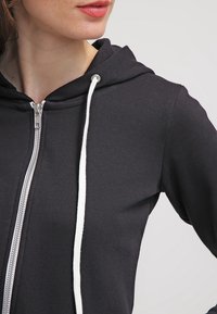 Sudadera con capucha negra con cremallera y cordón blanco, con textura acanalada, ajuste slim y detalle de cremallera metálica en la parte delantera.