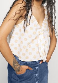 Žena v bezrukávovém bílém topu s žlutými puntíky a předním uzlem, v kombinaci s modrou džínovou sukní na knoflíky, má ruce v kapsách.