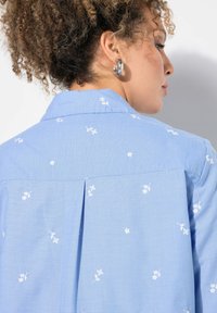 Camicia azzurro chiaro con ricami floreali bianchi, dotata di colletto classico e una piega sul retro per un dettaglio in più.