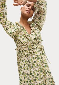 Blumendruckkleid mit Rüschendetails, in einer grünen und cremigen Farbpalette. Leichter Stoff mit fließendem Design und langen Ärmeln.
