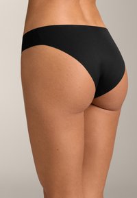 Žena oblečená v čiernych bezšvových bikinových nohavičkách, zobrazená zozadu na neutrálnom pozadí.