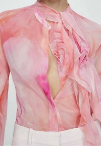 Blusa trasparente in rosa pallido, con colletto arricciato e bottoni. Il tessuto presenta un motivo acquerello con sfumature di rosa e corallo chiaro.