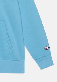 Light blue long-sleeve sweatshirt sleeve with ribbed cuff, kangaroo pocket edge, and small Champion logo embroidered near wrist.