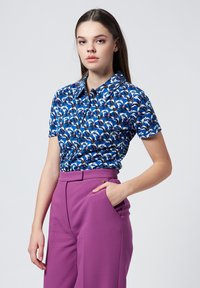 Short-sleeve shirt in blue fabric, featuring a multicolored bird print, paired with high-waisted purple trousers. Button-down collar detail visible.