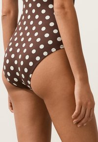 Costume da bagno marrone con grandi pois bianchi indossato da una persona vista dalla parte bassa della schiena fino a metà coscia, con un braccio rilassato.