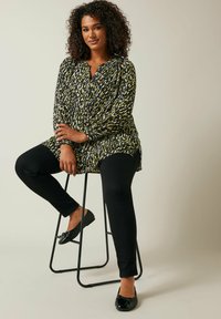 Blusa a maniche lunghe a fantasia nera e gialla, abbinata a pantaloni neri slim-fit e scarpe nere basse. Modella seduta su uno sgabello in metallo nero.