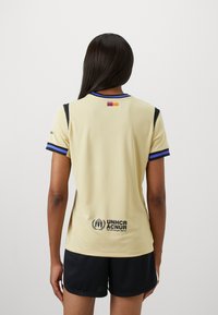 Camisa deportiva amarilla con acentos en azul y negro, que presenta un pequeño detalle de bandera en la parte superior de la espalda y el logo del ACNUR en el dobladillo inferior.