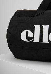 Svart sylinderformet veske laget av stoff, med en stor hvit logo. Den har en robust stropp og en teksturert overflate.