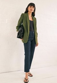 Blazer verde olivo sobre una blusa negra, combinado con jeans azul oscuro. Bolso negro trenzado, sandalias de tiras y denim de corte recto.