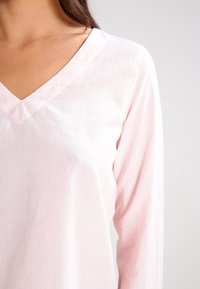 Haut à manches longues de couleur rose pâle avec un décolleté en V, présentant une texture subtile côtelée verticale. Tissu doux ; esthétique minimaliste.