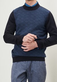 Uomo che indossa un maglione lavorato a maglia blu con maniche nere sopra una camicia blu con bottoni e pantaloni blu a trama.