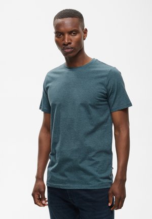 Junger Mann mit kurzen Haaren, der ein blau-grünes kurzärmeliges T-Shirt und dunkle Hosen trägt, steht vor einem schlichten hellen Hintergrund.