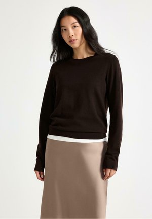 Donna con capelli neri indossa un maglione nero a maniche lunghe sopra una camicia bianca e una gonna beige, in piedi davanti a uno sfondo chiaro uniforme.