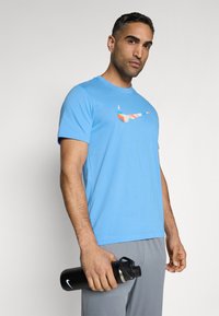 Camiseta de algodón azul con un logo de Nike colorido, acompañada de pantalones grises; sosteniendo una botella de agua negra con una tapa plateada.