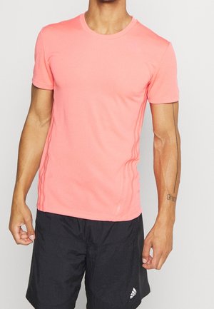 Mężczyzna w różowej, krótkodrukowanej sportowej koszulce i czarnych szortach Adidas, stojący z ramionami swobodnie opuszczonymi wzdłuż ciała.