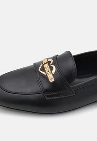 Mocassin en cuir noir avec logo "Love Moschino" doré et détail cœur sur la bride, avec surpiqûres visibles et finition lisse.